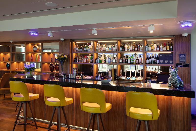 Tower Suites By Blue Orchid London Exterior photo The photo shows a modern bar area. In the foreground, there are several yellow bar stools lined up in front of a sleek black countertop. Behind the bar, there is a well-stocked shelf displaying a variety of bottles, including spirits and liqueurs, il