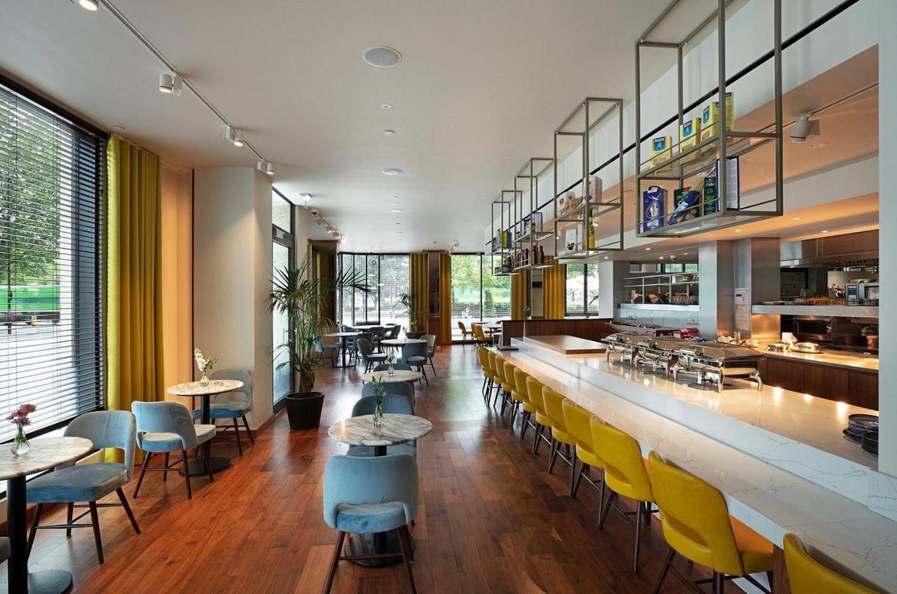 Tower Suites By Blue Orchid London Exterior photo The photo shows a modern restaurant interior. It features a spacious layout with large windows allowing natural light to flood the space. The decor includes a combination of wooden flooring and colorful furniture—blue and yellow chairs arranged aroun