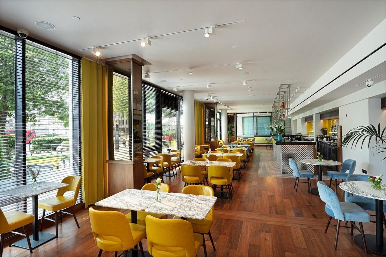 Tower Suites By Blue Orchid London Exterior photo The photo depicts a modern dining area with a bright and airy atmosphere. There are several tables with marble tops, accompanied by yellow chairs, creating a vibrant contrast with the wood flooring. The space features large windows that allow ample n