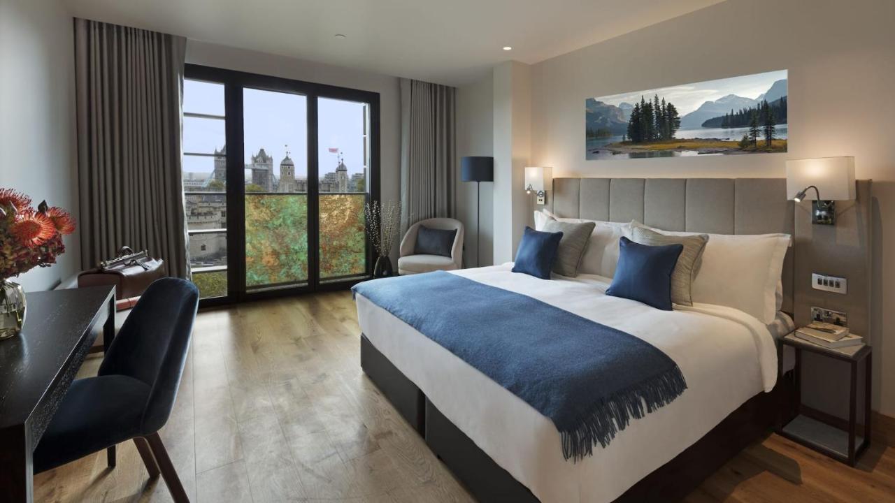 Tower Suites By Blue Orchid London Exterior photo The photo shows a modern hotel room that features a large bed with white linens and blue accent pillows. There’s a cozy throw blanket at the foot of the bed. The room is well-lit and includes a seating area with a chair and a small table. A large win