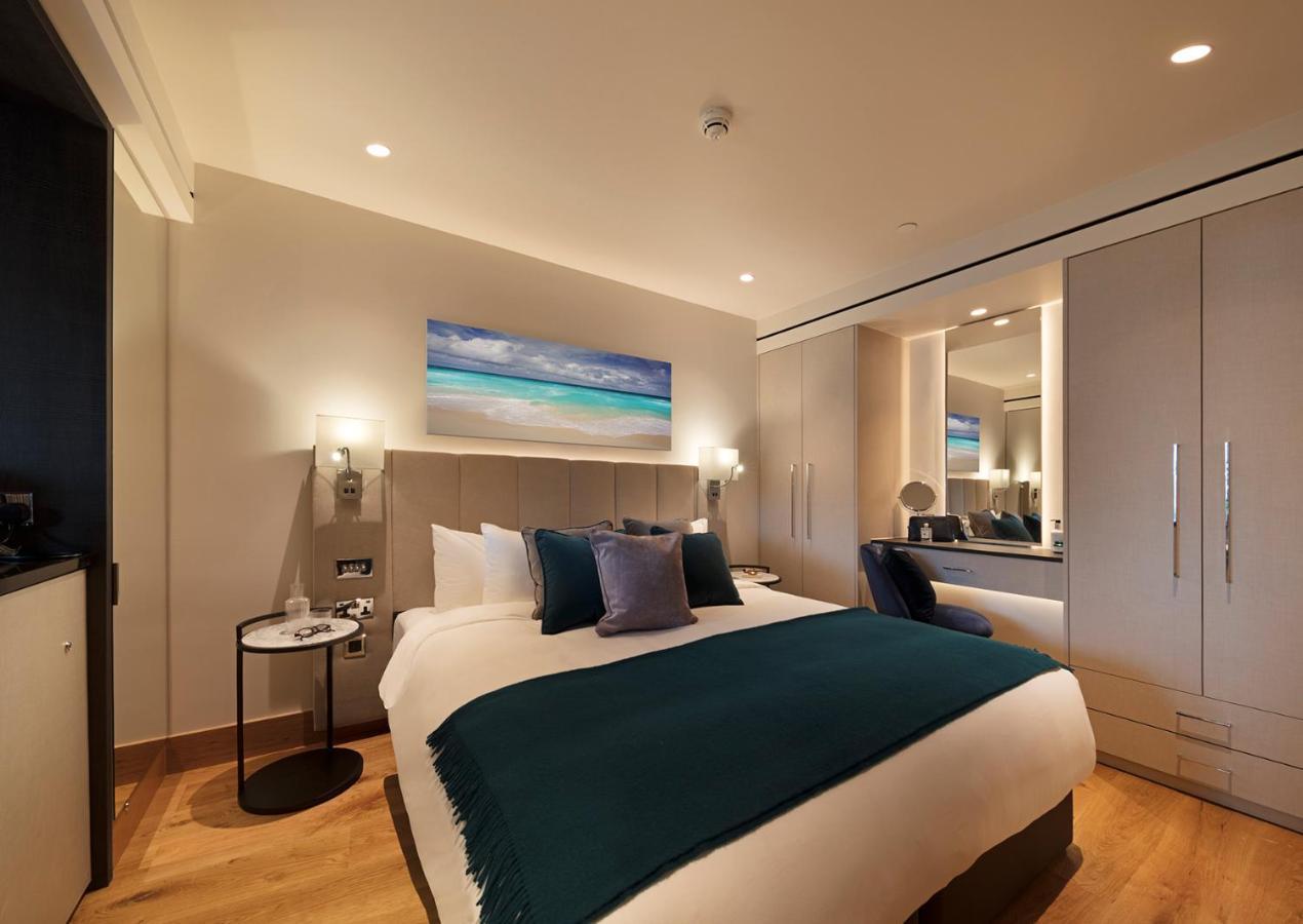 Tower Suites By Blue Orchid London Exterior photo The photo shows a modern hotel room featuring a double bed with a white duvet and a dark teal throw blanket. There are decorative pillows in shades of blue and purple on the bed. To one side, there's a round bedside table with a small lamp. A large m