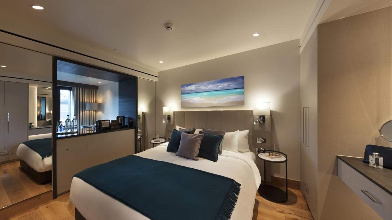 Tower Suites By Blue Orchid London Exterior photo The photo shows a modern hotel bedroom featuring a large bed with a light-colored headboard and a deep blue throw at the foot. The bed is adorned with multiple decorative pillows. On the wall above the bed, there is a colorful painting depicting a be