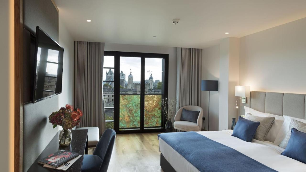 Tower Suites By Blue Orchid London Exterior photo The photo shows a modern hotel room featuring a large bed with blue and white bedding, a cozy armchair, and a small desk. There is a flat-screen television mounted on the wall across from the bed. The room has large glass doors leading to a balcony o