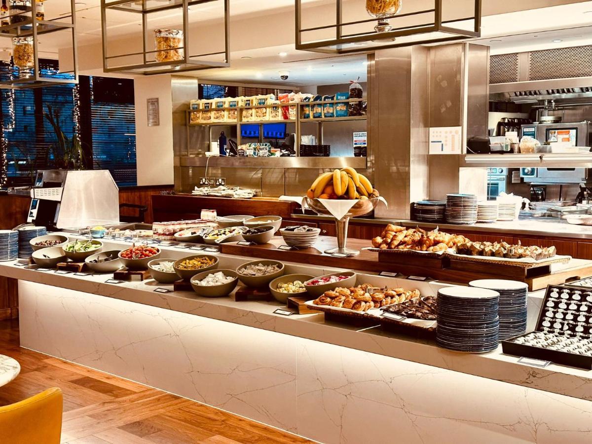 Tower Suites By Blue Orchid London Exterior photo The photo depicts a buffet-style setup, likely in a restaurant or a hotel dining area. In the foreground, there are multiple bowls and platters filled with a variety of food items, including salads, fruits, pastries, and other dishes. There is a long