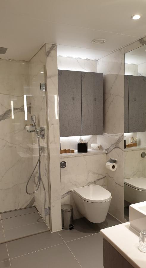 Tower Suites By Blue Orchid London Exterior photo The photo shows a modern bathroom design featuring elegant marble walls. On one side, there is a transparent shower enclosure with a showerhead and controls visible. Opposite the shower, there is a toilet, and above it are some sleek cabinets. The sp