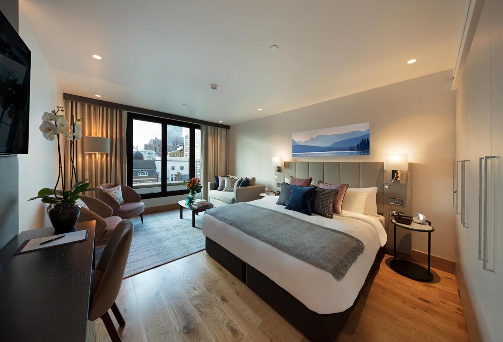 Tower Suites By Blue Orchid London Exterior photo The photo shows a modern hotel room. In the center, there is a large bed with stylish bedding and pillows in various shades. To one side, there are two comfortable chairs and a small table, creating a cozy seating area. A large window offers a view o